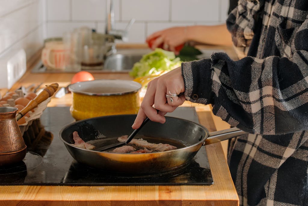 Person cooking bacon in a pan in a cozy kitchen setting. Fresh ingredients on the side.